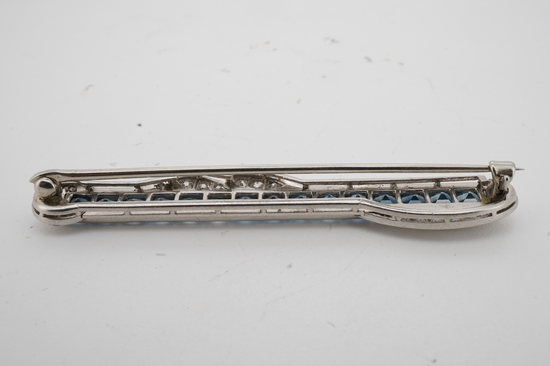 An Art Deco Aquamarine and diamond brooch, 1930s, of bar design, set with calibré-cut aquamarines, accented with foliate scrolls set with single-cut diamonds, length 5.5cm, white metal unmarked, gross weight 8 grams